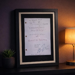 May include: A framed script page from "Inglourious Basterds" by Quentin Tarantino. The black frame surrounds a white page with handwritten text, including the title, director's name, and signatures. A small potted plant and a lamp are visible.
