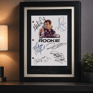 Peut inclure: Une couverture de script encadrée pour la série télévisée The Rookie, présentant une image centrale des acteurs et le titre de la série. Le papier blanc comporte plusieurs signatures à l'encre noire et bleue, sous un cadre noir.