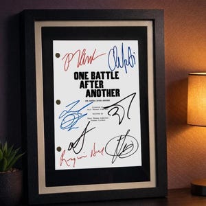 May include: Framed print featuring the title "ONE BATTLE AFTER ANOTHER" in black, with multiple signatures in red, blue, and black ink. The print is set against a white background, framed in black, and displayed on a dark surface.
