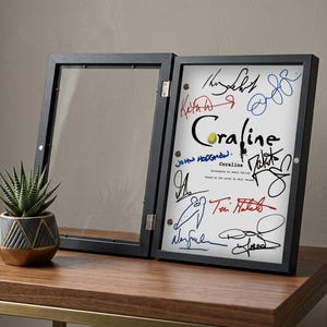 May include: A black framed display case, open to reveal a white print with colorful signatures and the title "Coraline." The frame is on a wooden table with a small potted succulent.
