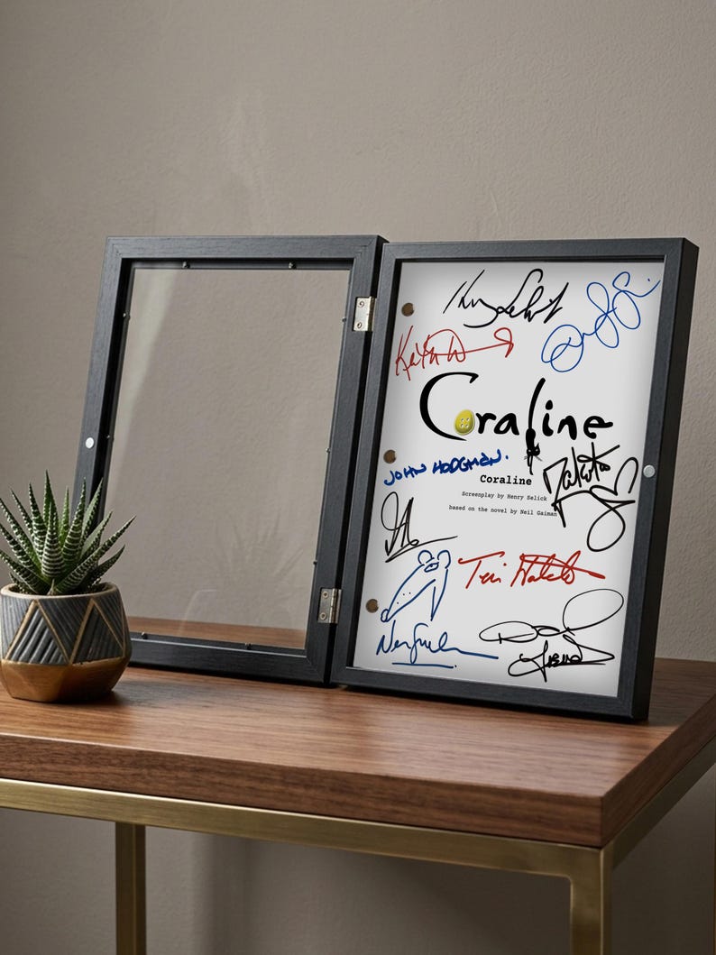 May include: A black framed display case, open to reveal a white print with colorful signatures and the title "Coraline." The frame is on a wooden table with a small potted succulent.