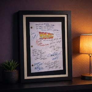 Puede incluir: Guion enmarcado de "Regreso al Futuro" con firmas. El guion se muestra en un marco negro con un passe-partout crema. El título "Regreso al Futuro" es visible, junto con notas manuscritas y firmas.