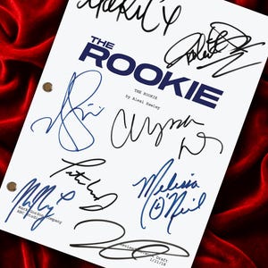 Puede incluir: Un guion de televisión blanco titulado The Rookie de Alexi Hawley, con múltiples firmas en tinta negra y azul en la página. El documento incluye tres orificios de encuadernación metálicos en el lado izquierdo y texto que indica un borrador.