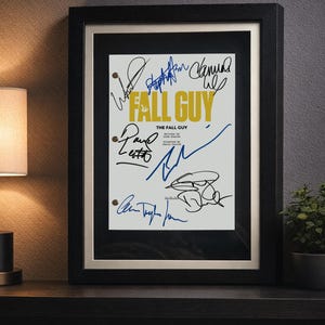 Może przedstawiać: Oprawiony plakat do filmu "Fall Guy" z podpisami. Tytuł jest żółty, z detalami filmu poniżej. Czarna rama ma złoty wewnętrzny brzeg. Plakat jest wystawiony na ciemnej półce.