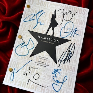 May include: A white script with the title "HAMILTON" and a silhouette of a person on a star. The script is signed in blue and black ink. The background is a deep red velvet.