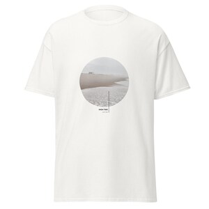 Könnte beinhalten: Ein weißes kurzärmeliges T-Shirt mit einer kreisförmigen Grafik auf der Brust. Die Grafik zeigt einen nebligen Strand mit Meereswellen und Sand. Unter dem Kreis steht der Text HIGH TIDE in schwarzen Großbuchstaben mit kleinerem Text darunter.