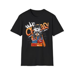 Könnte beinhalten: Schwarzes T-Shirt mit einem Cartoon-Katzen-Design, das eine goldene Kette trägt und einen Baseballschläger hält. Die Katze sitzt auf Kisten, mit orangefarbenem Graffiti "Who Cares?!".