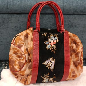 May include: A patterned handbag with red handles and accents. The bag features a black center panel with floral embroidery. The sides of the bag have a brown and tan pattern. The handles are a bright red color.
