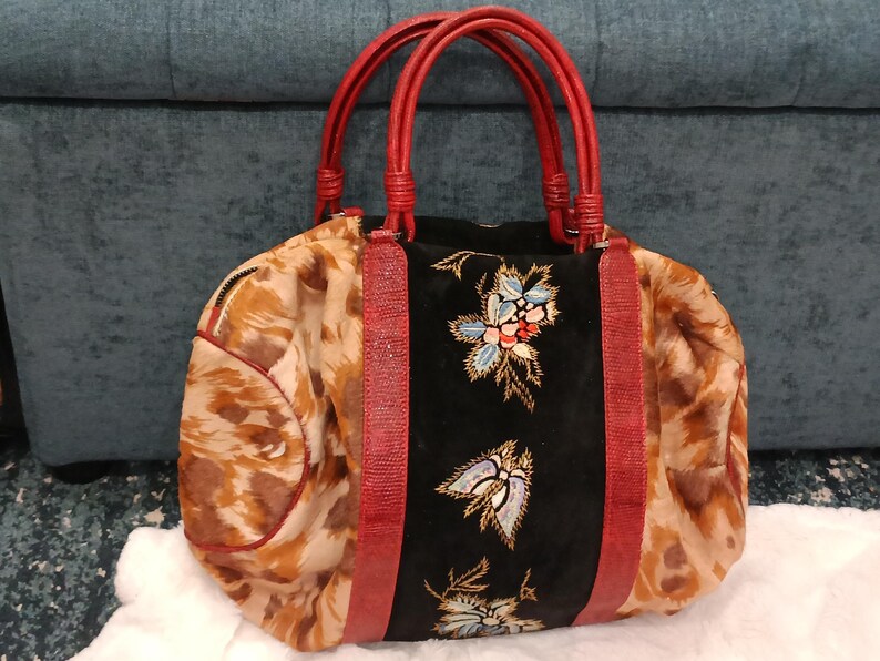 May include: A patterned handbag with red handles and accents. The bag features a black center panel with floral embroidery. The sides of the bag have a brown and tan pattern. The handles are a bright red color.