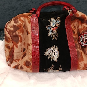 May include: A patterned handbag with a brown and tan animal print design. The bag features a black center panel with floral embroidery, red accents, and a red handle. A decorative charm hangs from the side.