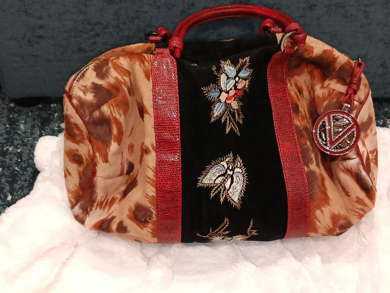 May include: A patterned handbag with a brown and tan animal print design. The bag features a black center panel with floral embroidery, red accents, and a red handle. A decorative charm hangs from the side.