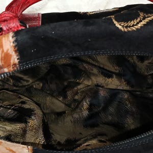 May include: A close-up of a handbag with a red handle and a black suede exterior. The bag features a patterned design in shades of brown and gold. The interior lining has a gold and black animal print.