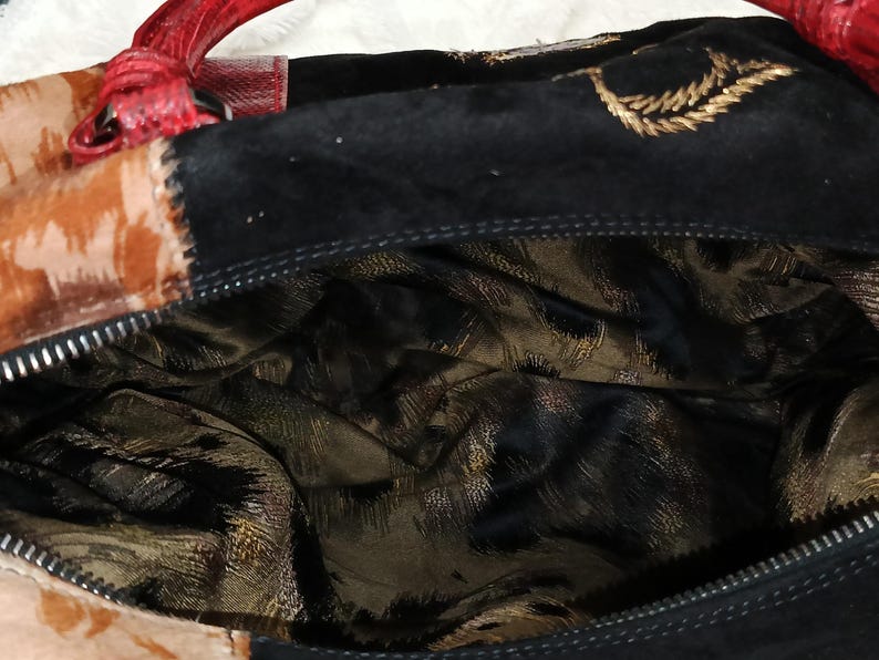 May include: A close-up of a handbag with a red handle and a black suede exterior. The bag features a patterned design in shades of brown and gold. The interior lining has a gold and black animal print.