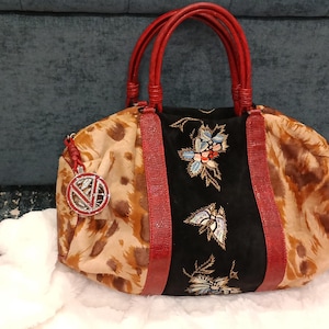 May include: A patterned handbag with red handles and accents. The bag features a black panel with floral embroidery and a decorative charm. The main body of the bag has a tan and brown animal print.