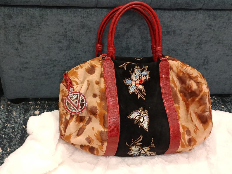 May include: A patterned handbag with red handles and accents. The bag features a black panel with floral embroidery and a decorative charm. The main body of the bag has a tan and brown animal print.