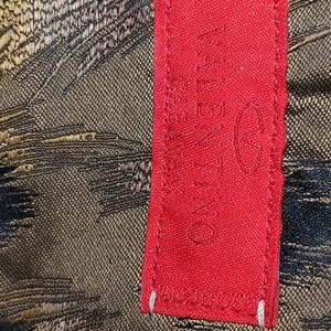 May include: Close-up of a red fabric tag with the brand name "VALENTINO" printed in white. The tag is next to a textured fabric with a pattern of black, gold, and brown threads.