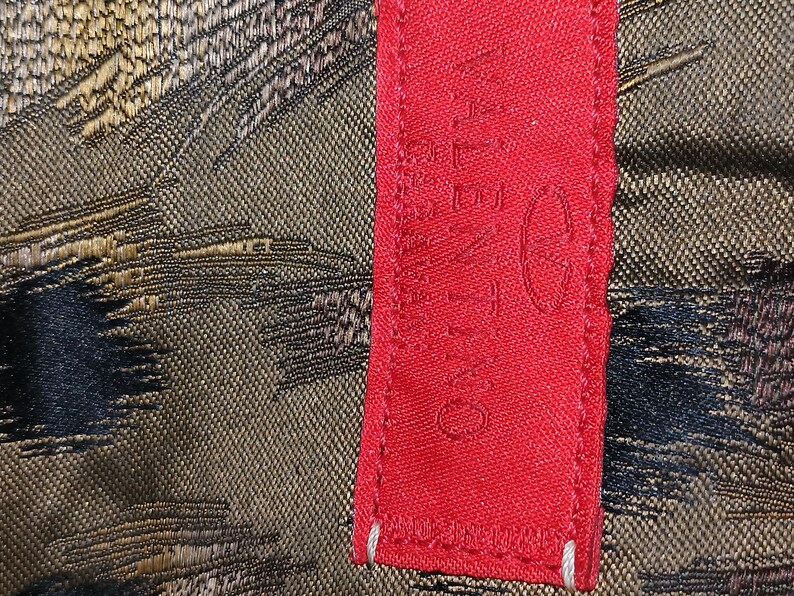 May include: Close-up of a red fabric tag with the brand name "VALENTINO" printed in white. The tag is next to a textured fabric with a pattern of black, gold, and brown threads.