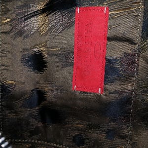 May include: Close-up of a textured fabric with a dark, abstract pattern in shades of brown, black, and gold. A bright red rectangular tag with the brand name "Valentino" is stitched onto the fabric with white thread.