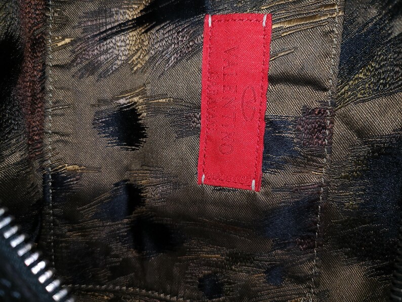 May include: Close-up of a textured fabric with a dark, abstract pattern in shades of brown, black, and gold. A bright red rectangular tag with the brand name "Valentino" is stitched onto the fabric with white thread.