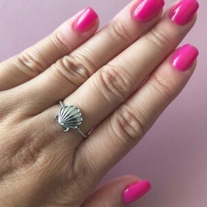 Pode incluir: Um anel de prata com um pingente de concha detalhado centrado numa banda de metal torcido. O anel é usado num dedo com verniz de unhas cor-de-rosa brilhante, destacando a textura metálica e as cristas distintas do design da concha.