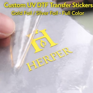 May include: Close-up of a clear sticker with gold foil lettering. The sticker features the word "HERPER" and a stylized "H" logo. The text "Custom UV DTF Transfer Stickers" is visible at the top, along with "Gold Foil / Silver Foil • Full Color".