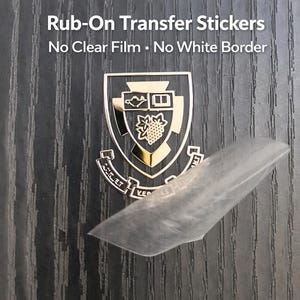 May include: A gold-colored rub-on transfer sticker with a shield design, featuring a grape cluster and other symbols. The text "Rub-On Transfer Stickers" and "No Clear Film &bull; No White Border" are visible.
