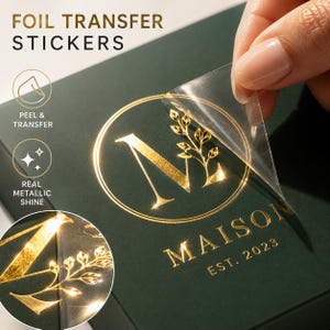 Op de afbeelding: Folie transfer stickers met een gouden metallic glans. De stickers worden van een donkergroen oppervlak verwijderd. Het ontwerp bevat het woord "MAISON" en het jaar "EST. 2023" in goud. De stickers zijn voor diverse toepassingen.