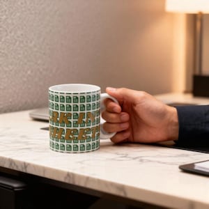 Puede incluir: Taza de cerámica blanca con un patrón verde repetido de iconos de archivos. Las palabras "TAKE IN SHEET" están impresas en letras doradas. La taza está sujeta por una mano.