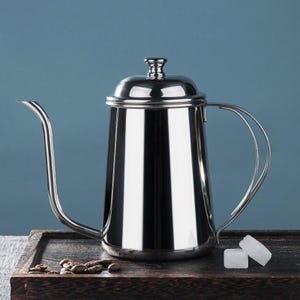 May include: A polished stainless steel gooseneck kettle with a long, curved spout and a handle. The kettle has a reflective surface and a lid with a small knob. It rests on a dark wooden tray with coffee beans and sugar cubes.