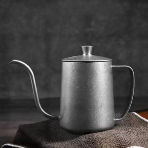 May include: A stainless steel gooseneck kettle with a long, curved spout and a small lid. The kettle has a handle and is designed for precise pouring, often used for pour-over coffee brewing. It sits on a brown cloth.