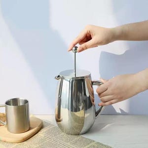 May include: A stainless steel French press coffee maker with a matching mug. The coffee press has a handle and a lid with a plunger. The mug is on a wooden coaster. The items are on a newspaper.