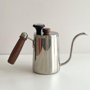 Puede incluir: Una tetera de cuello de cisne de acero inoxidable con asa y tapa de madera. La tetera tiene un pico largo y curvado para verter con precisión. Un termómetro está en la parte superior de la tapa. El asa es de madera marrón oscuro.
