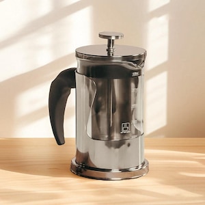 May include: A stainless steel French press coffee maker with a black handle. The clear glass carafe has measurement markings in milliliters. The lid and base are made of shiny metal. The coffee maker sits on a wooden surface.