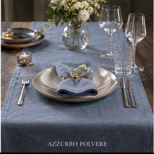 May include: A blue linen table runner set with a place setting featuring two stacked ceramic plates, a blue cloth napkin held by a gold ring, a silver fork, two silver knives, two clear wine glasses, a textured glass, and a vase with white flowers.