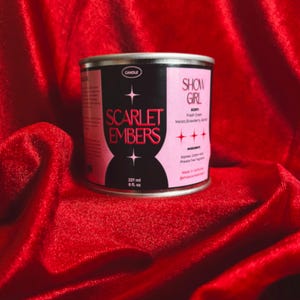 Può includere: Un barattolo di candela rotondo con un'etichetta rosa e nera. L'etichetta recita "SCARLET EMBERS" e "SHOW GIRL". La candela contiene 237 ml di prodotto. La candela è su una superficie di velluto rosso.