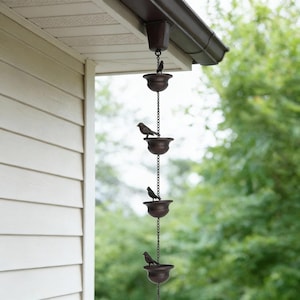 May include: A decorative rain chain with a series of bronze-colored bowls, each adorned with a small bird figurine. The chain hangs from a brown gutter, with a backdrop of a light-colored house and green foliage.