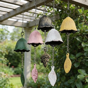 May include: A collection of colorful wind chimes hanging from a wooden pergola. The chimes are bell-shaped with leaf-shaped pendants, in shades of green, pink, white, yellow, and black. The background features lush greenery.