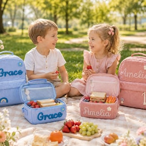 May include: A light blue backpack and matching lunch box with the name Grace embroidered in blue, alongside a dusty pink backpack and matching lunch box with the name Amelia embroidered in white, both featuring floral and rainbow accents.