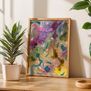 May include: Abstract watercolor art print in a wooden frame. The artwork features a colorful blend of pink, purple, blue, and yellow hues. The framed art is displayed on a wooden table, with a potted plant on the left and another on the right.