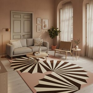 May include: A living room with a grey sofa, a round wooden coffee table, and a geometric rug with brown and cream fan-like patterns on a pink background. The room has light-coloured walls, a wooden chair, and a potted plant.