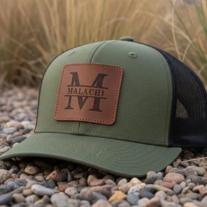 May include: An olive green and black trucker cap with a brown leather patch. The patch displays the name "MALACHI" and the initial "M" in a bold, stylized font. The cap is resting on a bed of small pebbles.