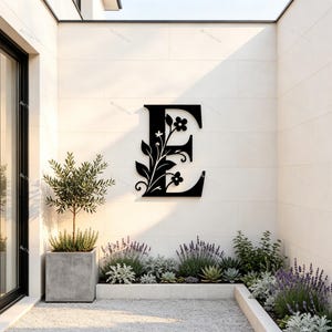 Könnte beinhalten: Ein großer, schwarzer, an der Wand montierter Buchstabe E aus Metall mit einem integrierten dekorativen Design aus Blumenstielen, Blättern und Blüten, die an der linken Seite des Buchstabens befestigt sind und einen Silhouetteneffekt erzeugen.