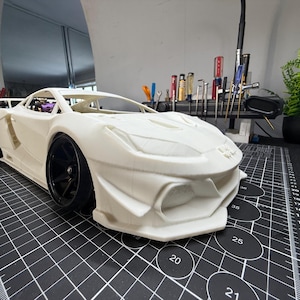 May include: A white, detailed model sports car with black wheels sits on a black cutting mat. The car has a low profile, aerodynamic design, and a prominent front splitter. Tools and a small plant are visible in the background.