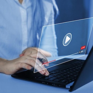 May include: A person using a black laptop with a video streaming interface overlaid on the screen. The interface features a play button, a live indicator, and a progress bar. The person is wearing a light blue button-down shirt.