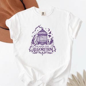 May include: White t-shirt with a purple graphic featuring a birdcage, flames, stars, and the text "ESCAPED THE ALGORITHM". The design is centered on the front of the shirt.
