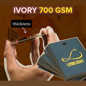 May include: A stack of square dark blue hang tags with a gold logo and the text LOWLIGHT, alongside a single tag held up to show its ivory 700 GSM thickness. The tags feature a small hole at the top for attachment, showcasing a sturdy paper quality.