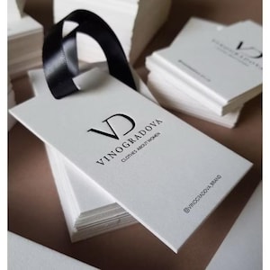May include: A white rectangular paper hang tag with a black ribbon loop. The tag features the black printed text VD VINOGRADOVA CLOTHES ABOUT WOMEN and the social media handle @VINOGRADOVA_BRAND. Multiple identical tags are stacked underneath.