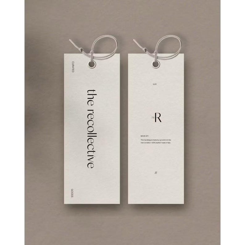 May include: Two rectangular off-white paper tags with rounded corners and a hole at the top, each threaded with a thin string. The left tag features the text the recollective, while the right tag displays the letter R and additional small text.