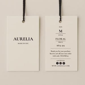 May include: Two rectangular off-white paper hang tags with black string loops. The left tag displays AURELIA and MADE IN NYC. The right tag lists SIZE M, STYLE FLORAL, and includes text about a purchase, a discount code, social media icons, and a website.