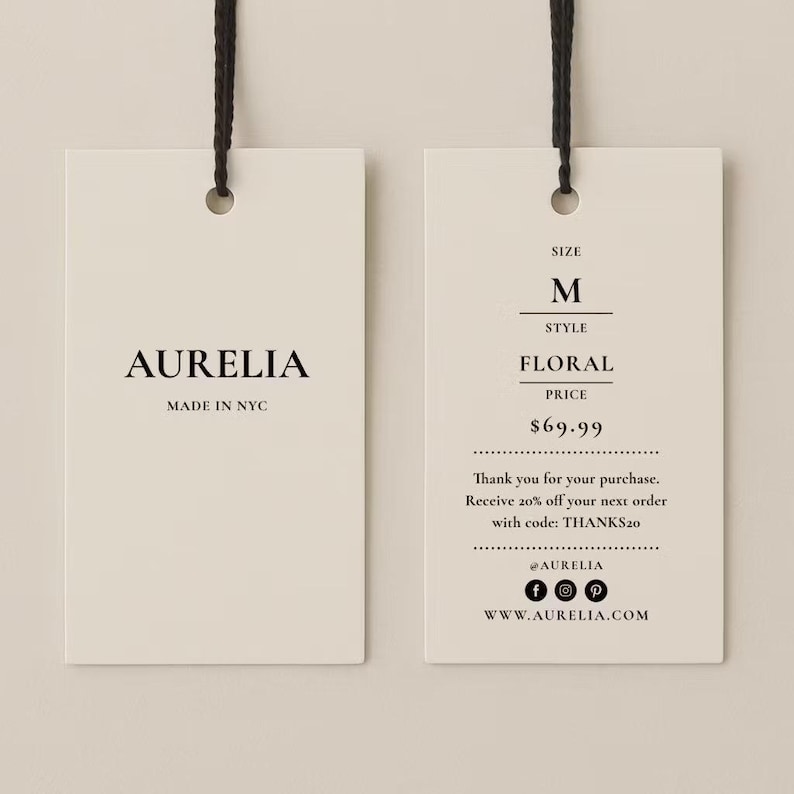 May include: Two rectangular off-white paper hang tags with black string loops. The left tag displays AURELIA and MADE IN NYC. The right tag lists SIZE M, STYLE FLORAL, and includes text about a purchase, a discount code, social media icons, and a website.